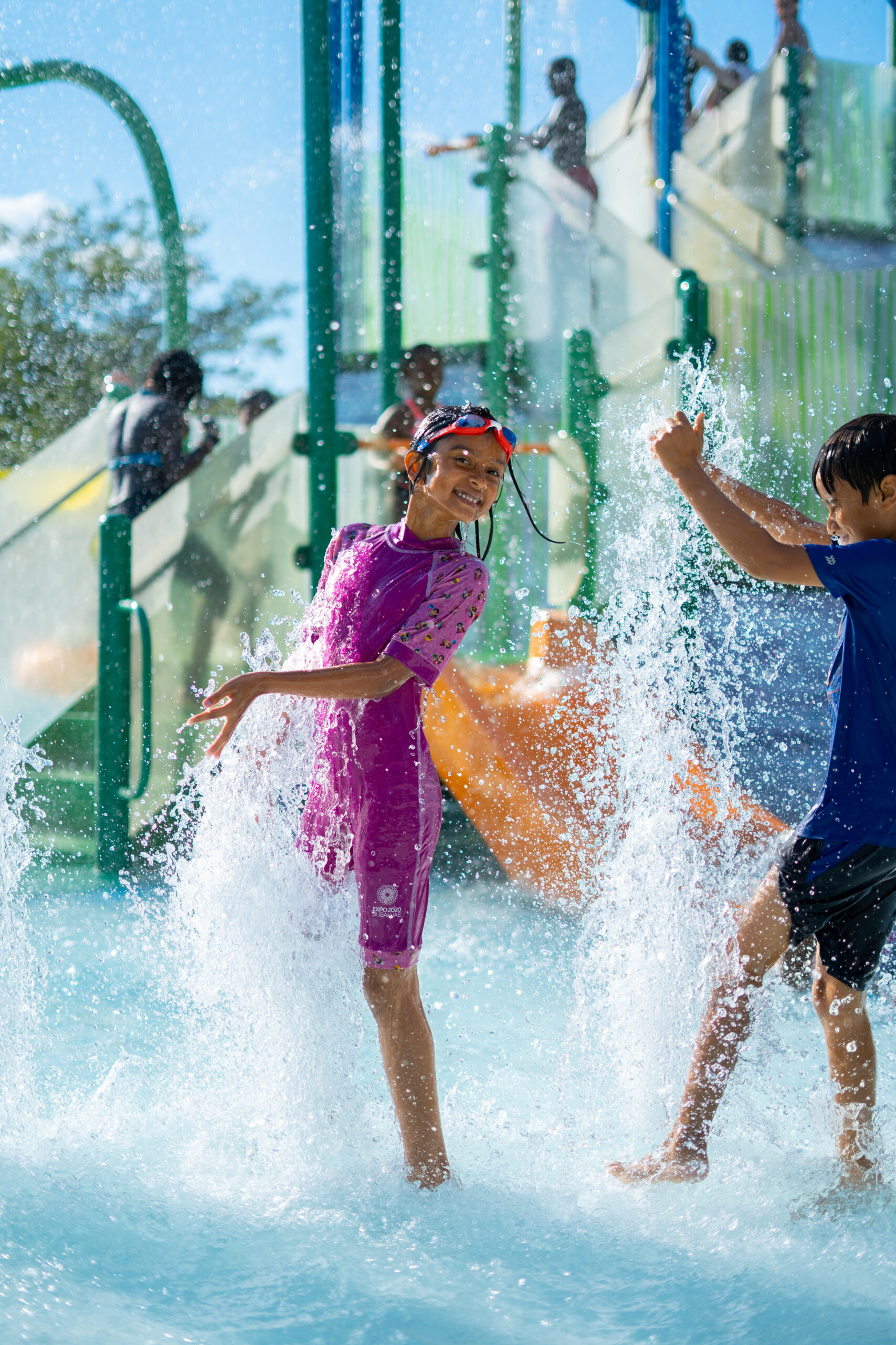 Children smile at the camera and splash in an upward spray feature in the activity pool at The Waterpark.