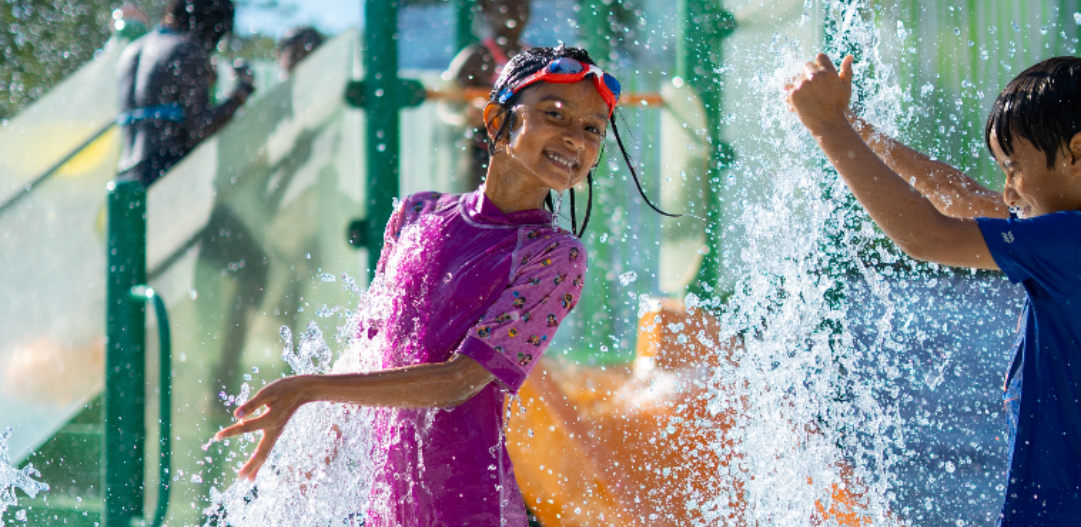Children smile at the camera and splay in a spray-up feature at the Activity Pool at The Waterpark.