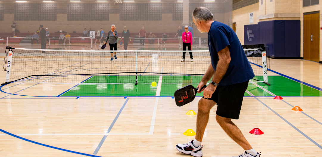 Pickleball players serve the ball across the net in the MCC gymnasium.