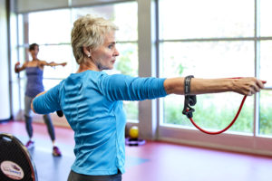 MCC group fitness class for seniors, with woman using a stretch band pulled back like an arrow in front of a wall of windows.