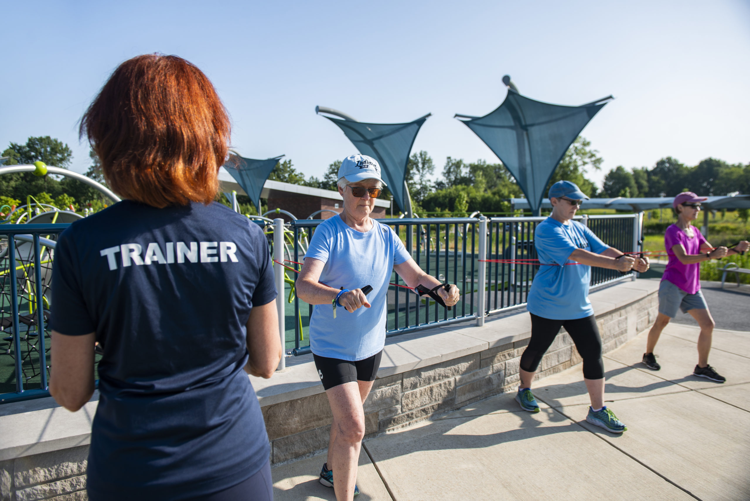Trainer works with 3 training clients during a group fitness class. Participants use stretch bands along the wall at Central Park.