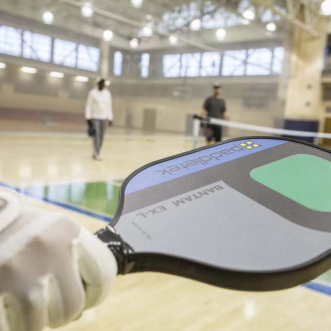 Pickelball paddle in the foreground with a view of the pickleball courts set up in the gym.