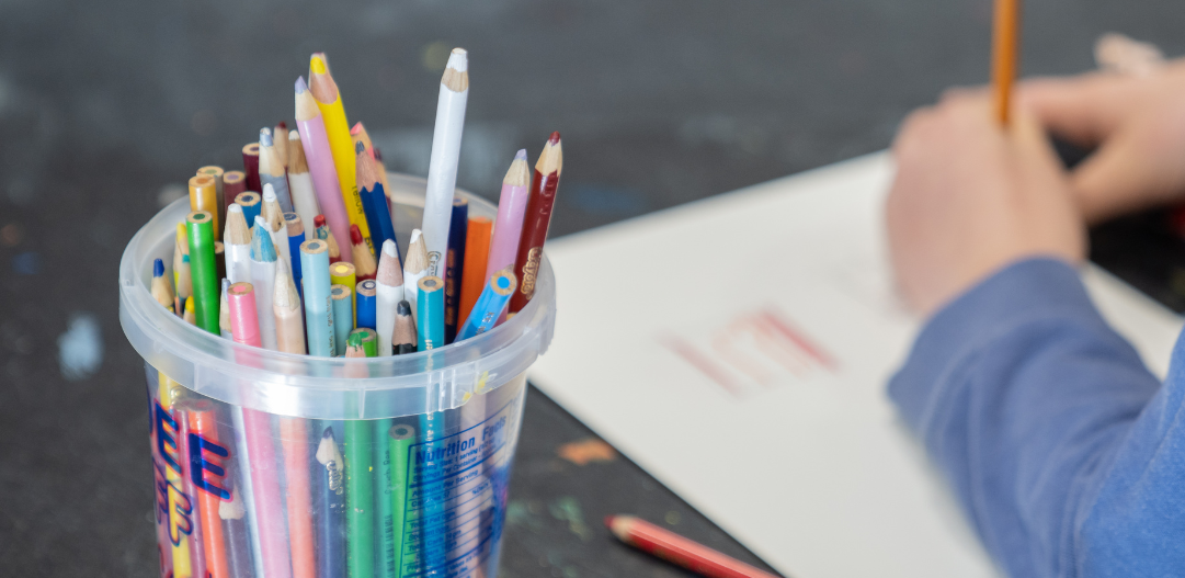 A bucket of colored pencils sits beside a child with a pencil in hand and paper creating a work of art in an art class at the Monon Community Center.