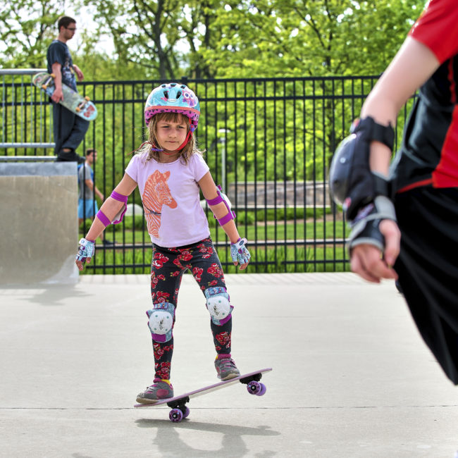 Young girl rolls across the screen on her skateboard with her pink and teal helmet, bright flowery outfit and kneepads.
