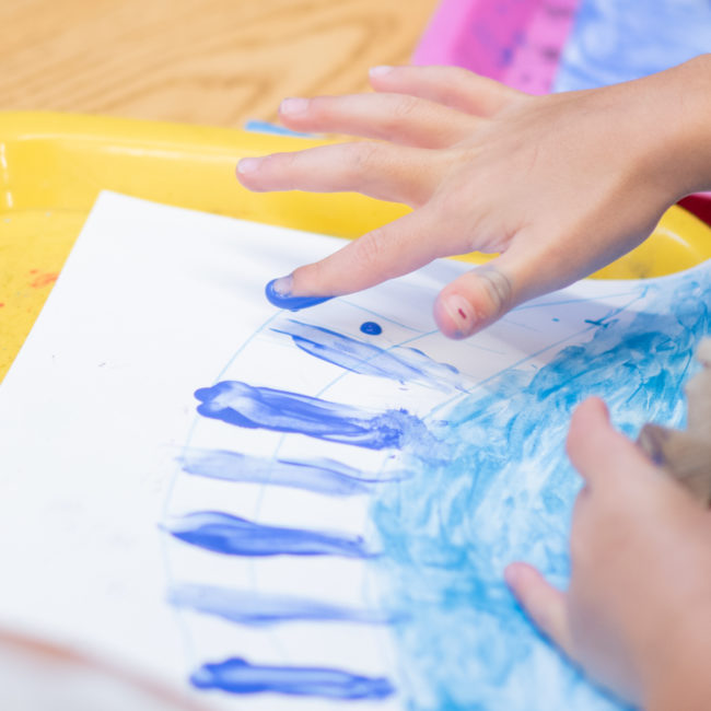 Child finger paints lines along a white paper with bright blue finger paint, set against a yellow paint tray.