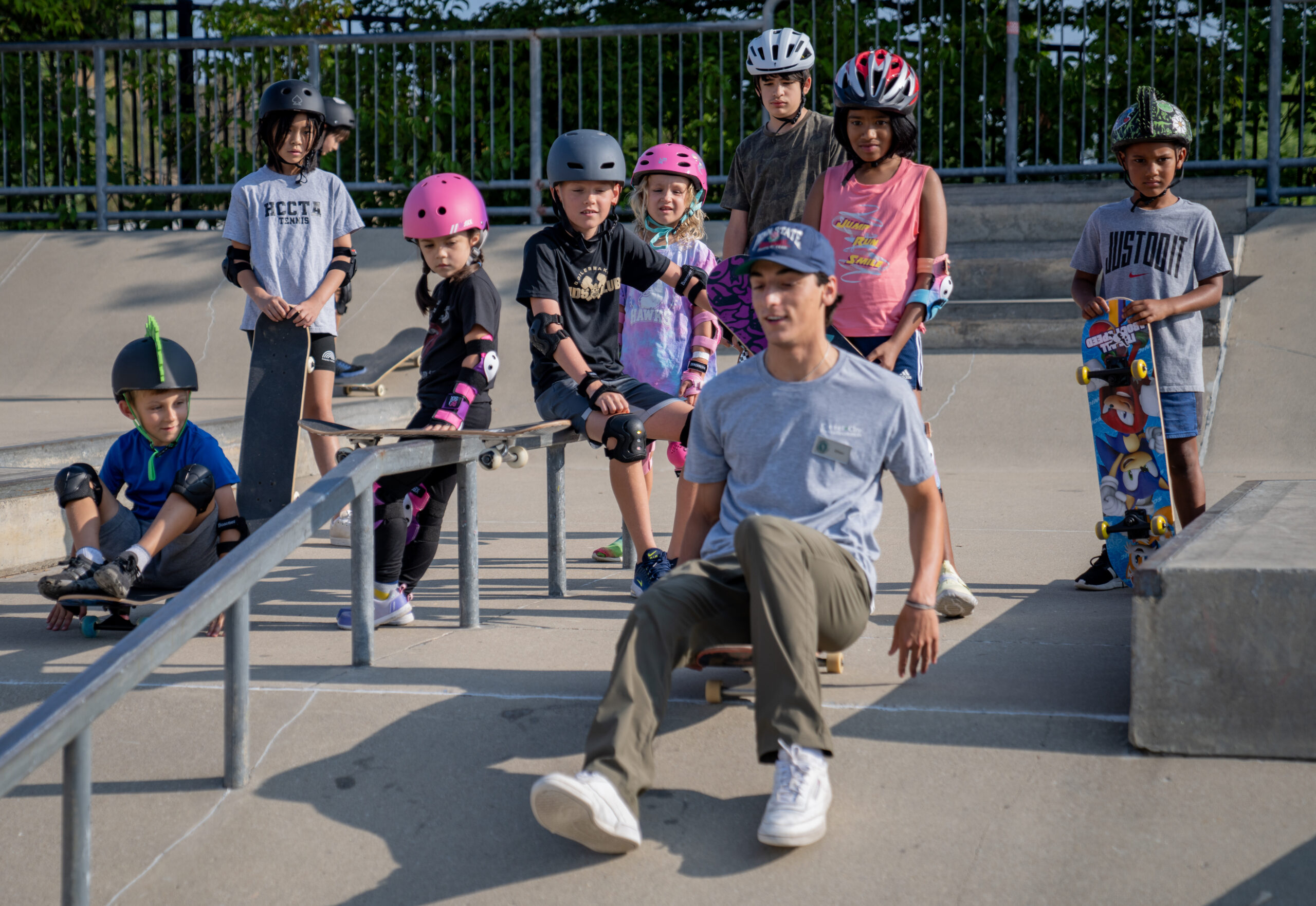 Skateboarding instructor shows kids how to get used to the feeling of the skateboard going downhill by sitting on the board and going down the side of the ramp on your bottom at the Skatepark.