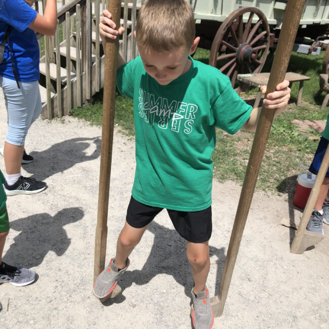 Student tries to walk on stilts at Conner Prairie during Camp En Route.
