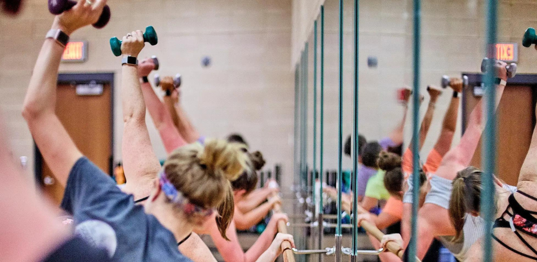 Barre class in a group fitness studio with mirrors and weights.