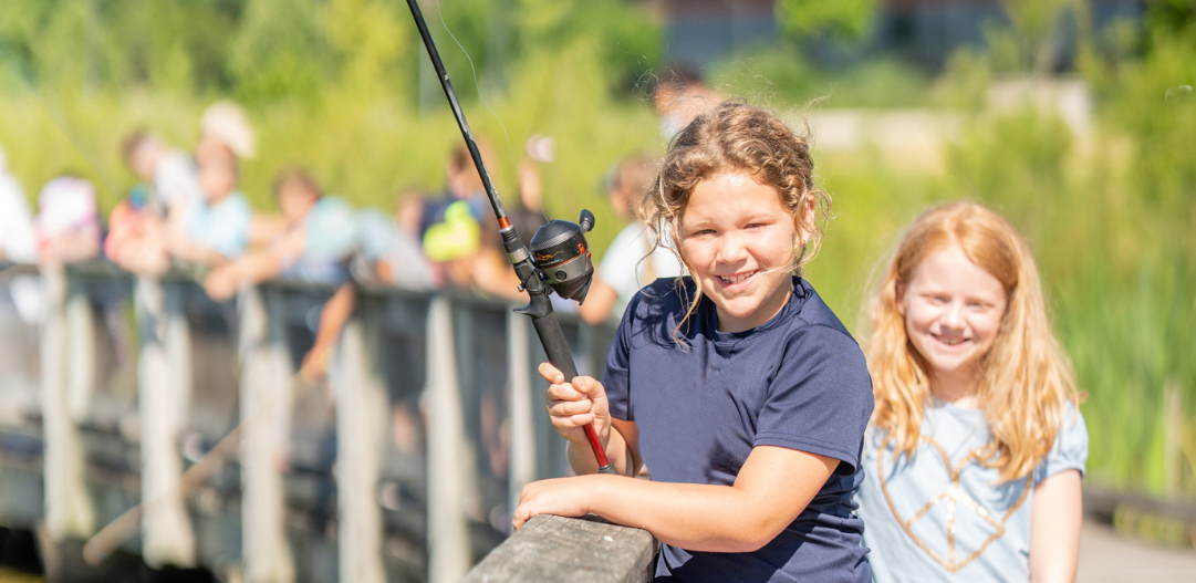 2 girls in the foreground smiling with their fishing poles on a bright sunny day at the docks at West Park for Outdoor Explorer camp.