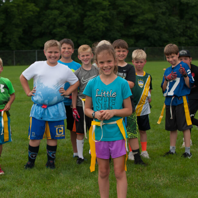 Participants stand before camera in the grass for a pose after their final day of flag football for Kids at Play summer camp.