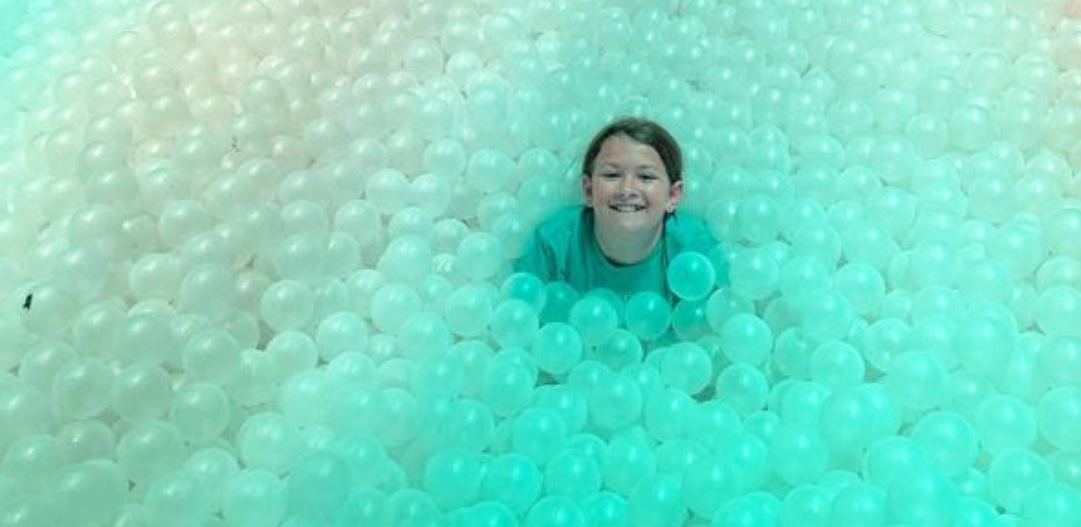 Child smiles up at he camera while being immersed all the way up to their neck in plastic balls in a ball pit.