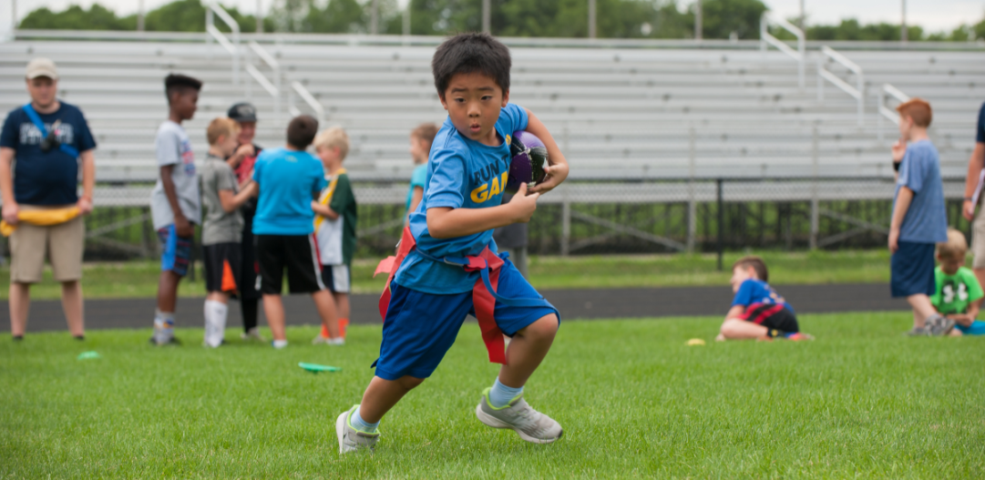 Child runs with the football, bleachers and field in the background, during a flag football game at Kids at Play camp.