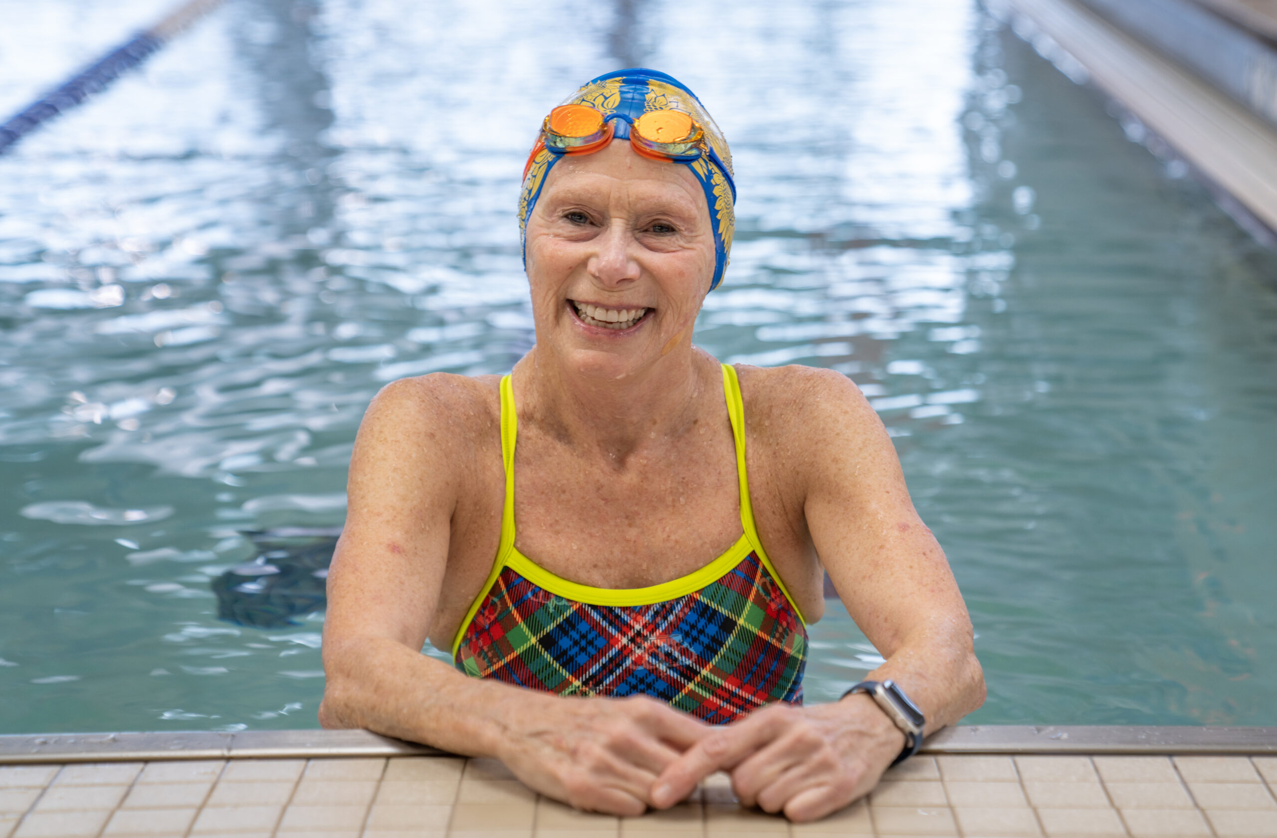 MCC member Robin W. smiles at the edge of the indoor lap pool with goggles and a swimcap.